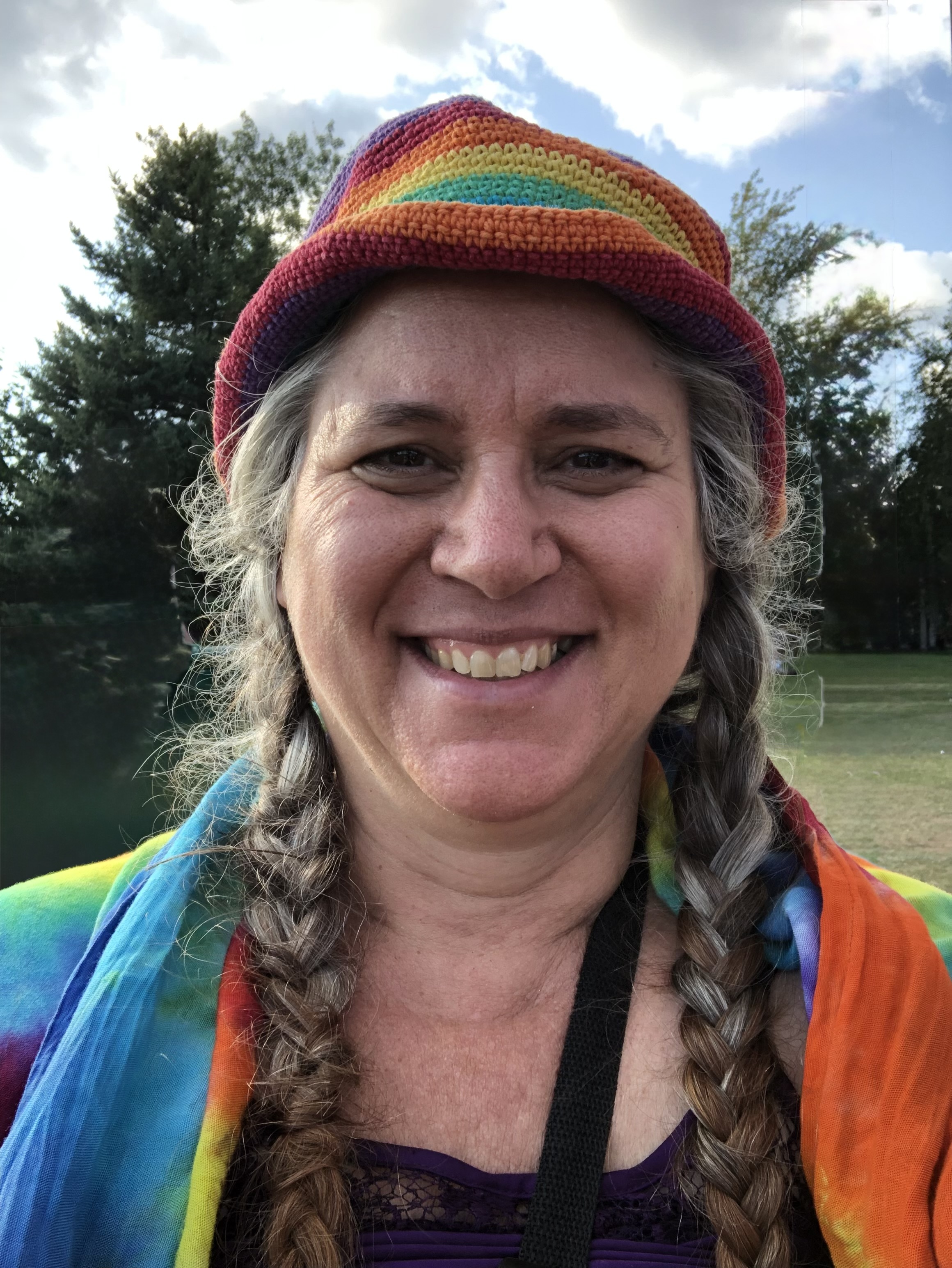 Diane smiles at the camera wearing a rainbow hat and shawl with a blue sky and trees in the background.